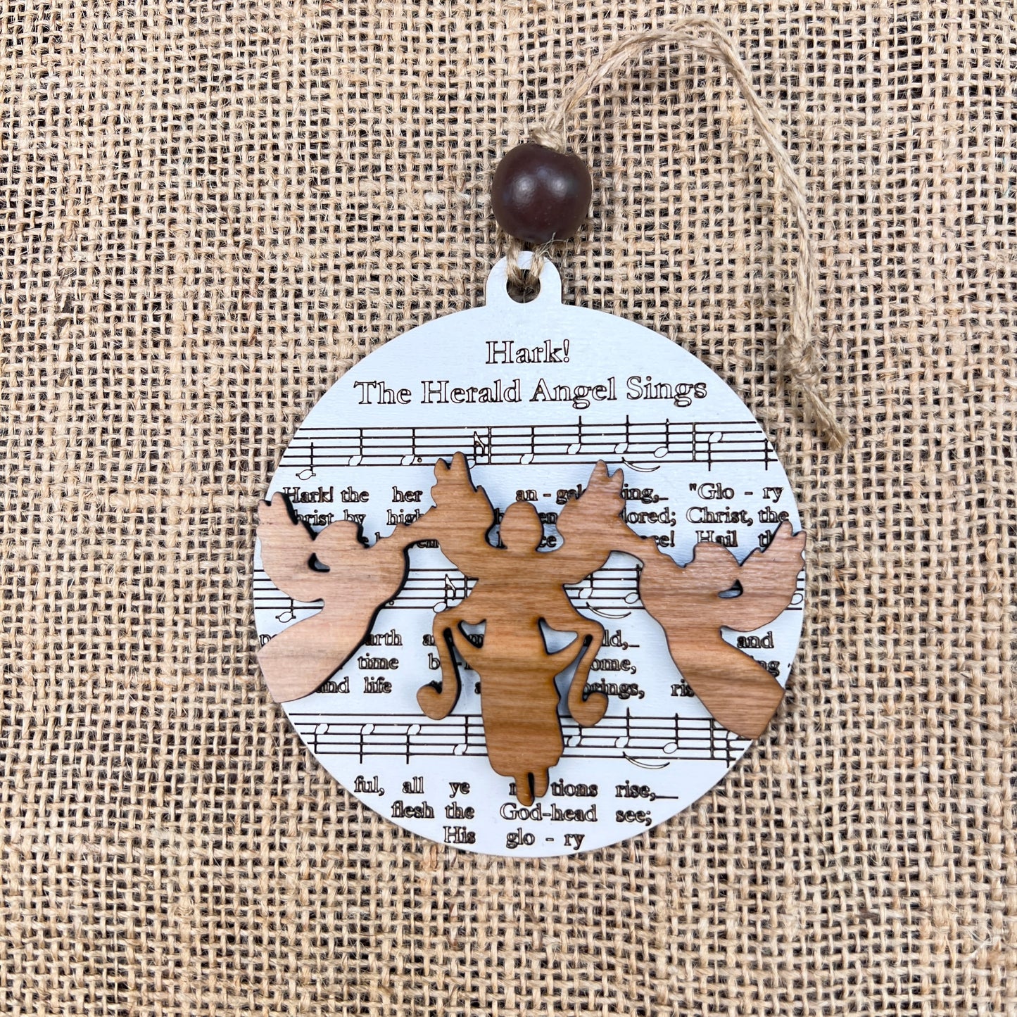 This round ornament from Janet's Craft Corner exudes rustic charm with cutout wooden angels set against sheet music for "Hark! The Herald Angels Sing." Perfect for music lovers, the Sheet Music Ornaments feature a burlap background with a loop and bead for easy hanging.