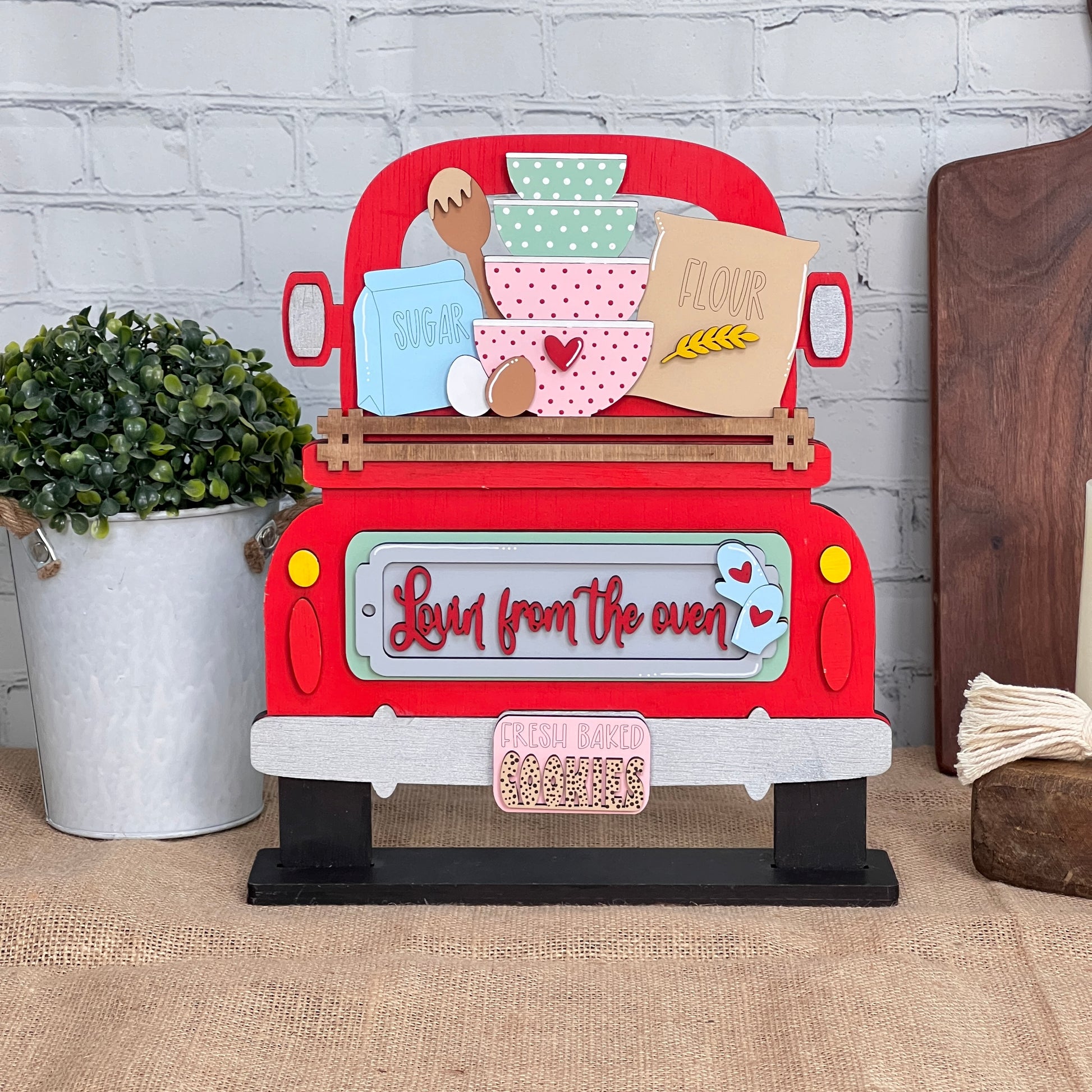 This DIY craft kit from Janet's Craft Corner, named "Lovin from the Oven Interchangeable Insert," features a hand-painted decorative sign shaped like a red truck. It is adorned with baking-themed items such as a mixing bowl, flour, and sugar. Ideal for home decor, the sign reads "Love from the oven" and "Fresh baked cookies," and comes with interchangeable inserts and a potted plant.