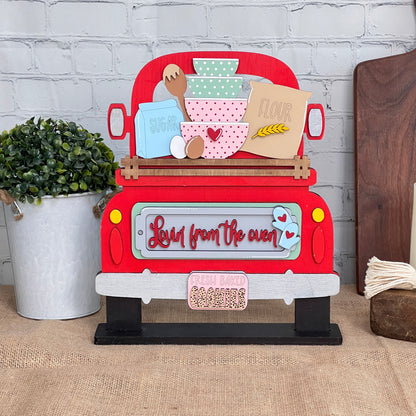 This DIY craft kit from Janet's Craft Corner, named "Lovin from the Oven Interchangeable Insert," features a hand-painted decorative sign shaped like a red truck. It is adorned with baking-themed items such as a mixing bowl, flour, and sugar. Ideal for home decor, the sign reads "Love from the oven" and "Fresh baked cookies," and comes with interchangeable inserts and a potted plant.