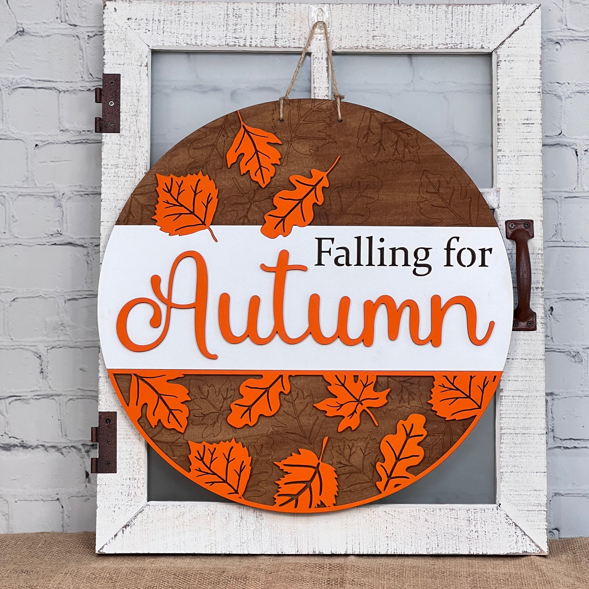 The Falling for Autumn Round DIY Home Decor Kit by Janet's Craft Corner features a charming round wooden sign adorned with orange leaves and the phrase "Falling for Autumn." This delightful piece hangs elegantly on a rustic white frame with a brown handle, making it an ideal personalized home décor addition that pairs beautifully with a white brick wall backdrop.