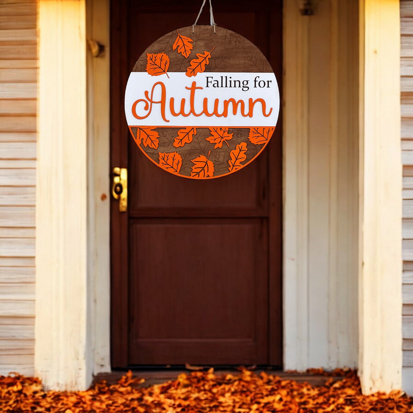 The Falling for Autumn Round DIY kit from Janet's Craft Corner features a hand-painted sign with orange leaves, perfect for enhancing home décor with a cozy touch against white siding and leaf-covered ground.