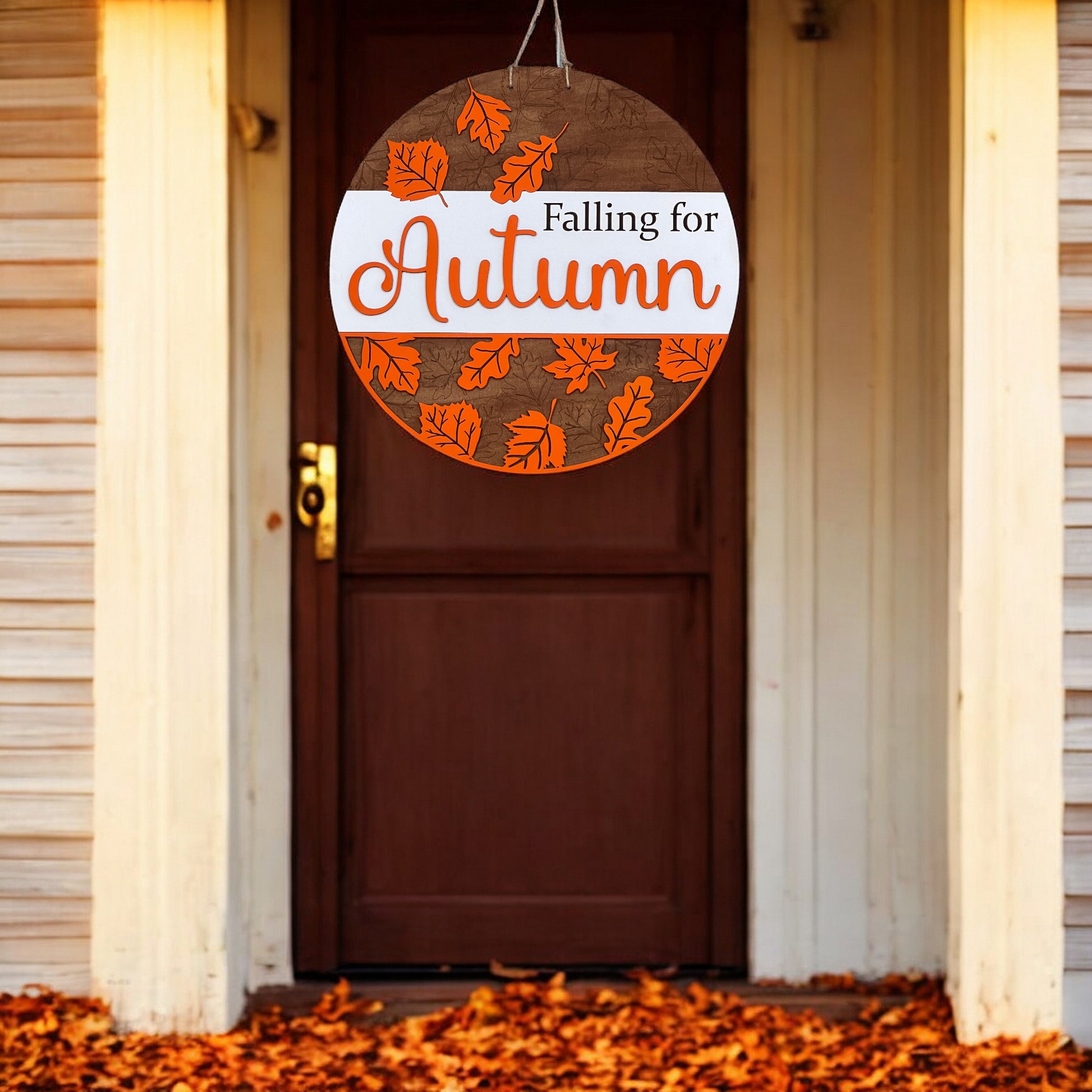 The Falling for Autumn Round DIY kit from Janet's Craft Corner features a hand-painted sign with orange leaves, perfect for enhancing home décor with a cozy touch against white siding and leaf-covered ground.