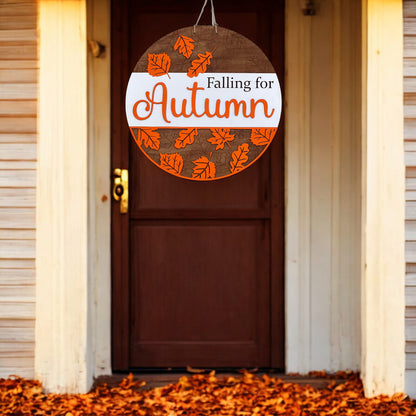 The Falling for Autumn Round DIY kit from Janet's Craft Corner features a hand-painted sign with orange leaves, perfect for enhancing home décor with a cozy touch against white siding and leaf-covered ground.