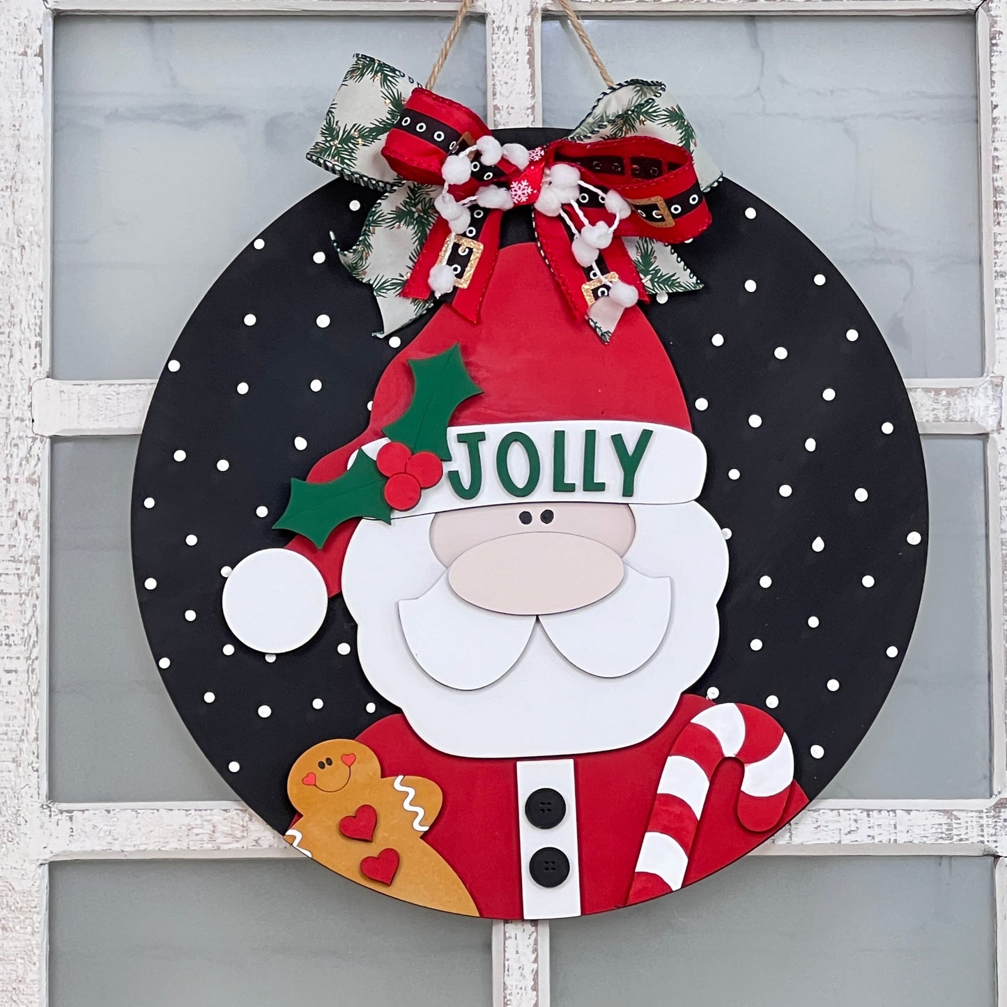 The Jolly Santa Round Sign from Janet's Craft Corner is a delightful holiday DIY home decor kit. It features Santa Claus wearing a "Jolly" hat set against a black polka dot backdrop, holding a gingerbread man and candy cane. A festive ribbon adorned with holly and bells completes this charming hand-painted decoration.