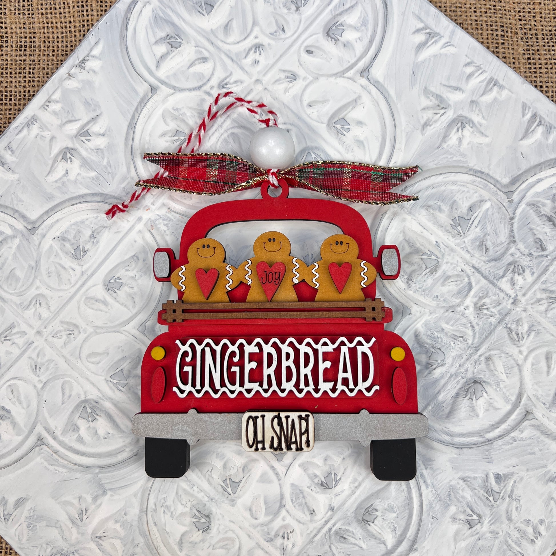 The Antique Truck Ornament from Janet's Craft Corner is a charming addition to your Christmas décor collection, featuring a red pickup loaded with gingerbread figures. The tailgate reads "GINGERBREAD" with "OH SNAP!" displayed below. A plaid bow atop the ornament completes the festive look, set against a textured white background.