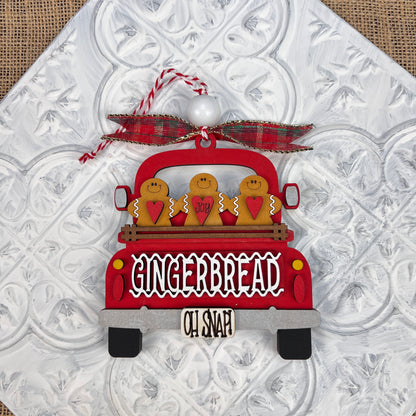 The Antique Truck Ornament from Janet's Craft Corner is a charming addition to your Christmas décor collection, featuring a red pickup loaded with gingerbread figures. The tailgate reads "GINGERBREAD" with "OH SNAP!" displayed below. A plaid bow atop the ornament completes the festive look, set against a textured white background.