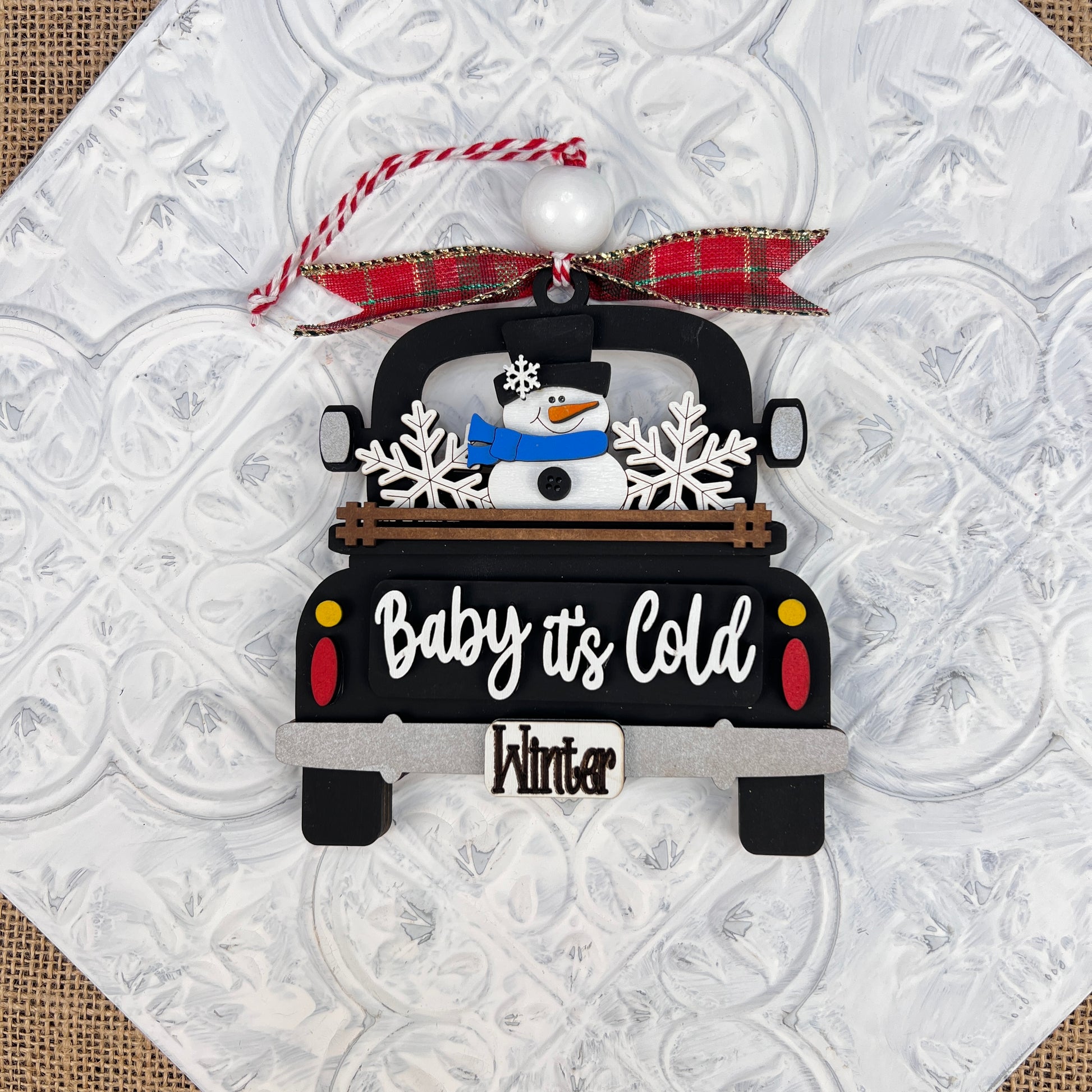 This DIY kit from Janet's Craft Corner features an Antique Truck Ornament, complete with a charming snowman and snowflakes in the back. The black truck is adorned with the phrase "Baby it's Cold," while the license plate reads "Winter," all accented by a red plaid ribbon. It's an ideal addition to your Christmas décor collection.