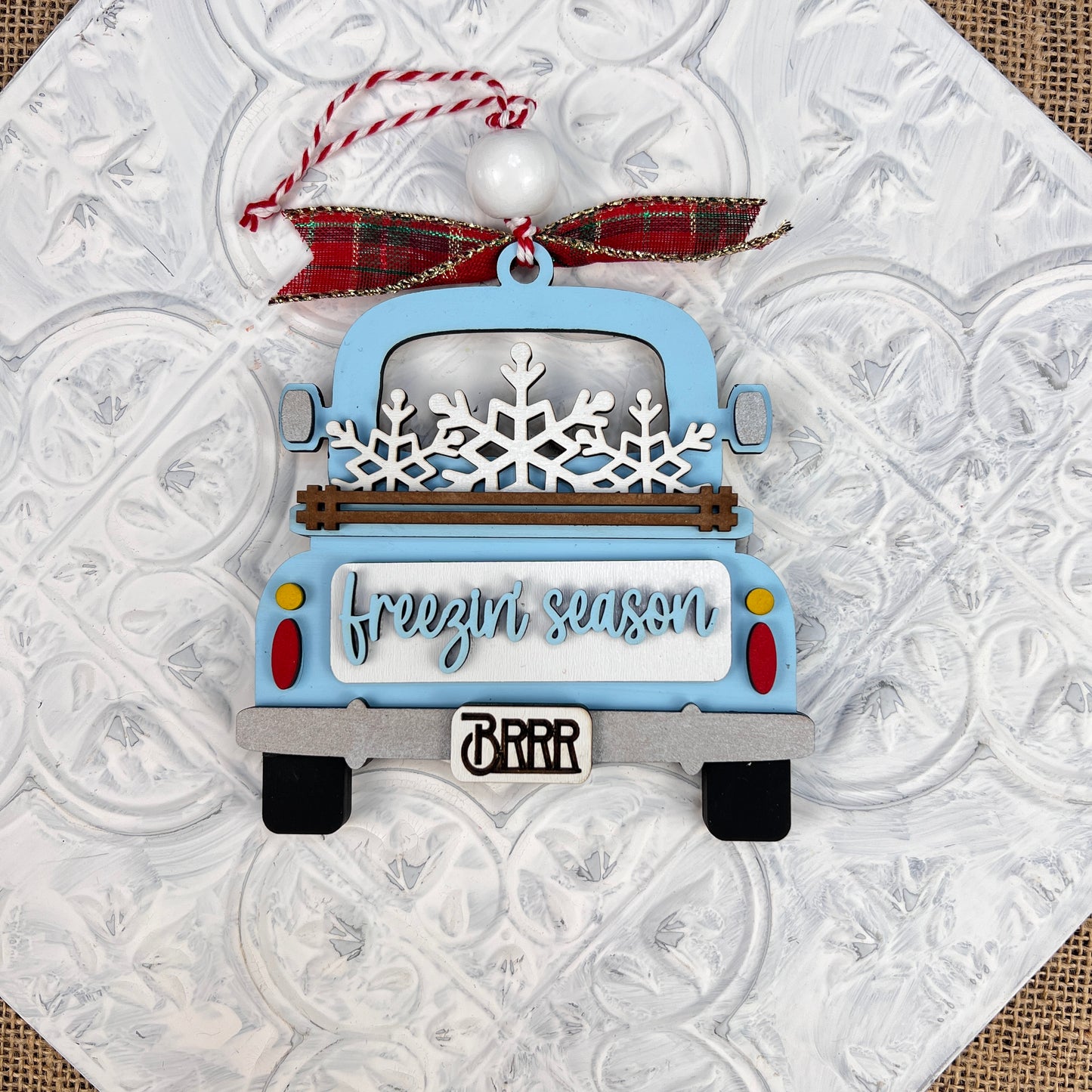 The Janet's Craft Corner Antique Truck Ornament features a charming blue truck adorned with snowflakes in the back, accompanied by the phrase "freezin' season" on the tailgate. It hangs from a festive red and green plaid ribbon against a textured white background, making it an ideal addition to your Christmas décor.