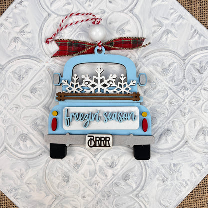 The Janet's Craft Corner Antique Truck Ornament features a charming blue truck adorned with snowflakes in the back, accompanied by the phrase "freezin' season" on the tailgate. It hangs from a festive red and green plaid ribbon against a textured white background, making it an ideal addition to your Christmas décor.