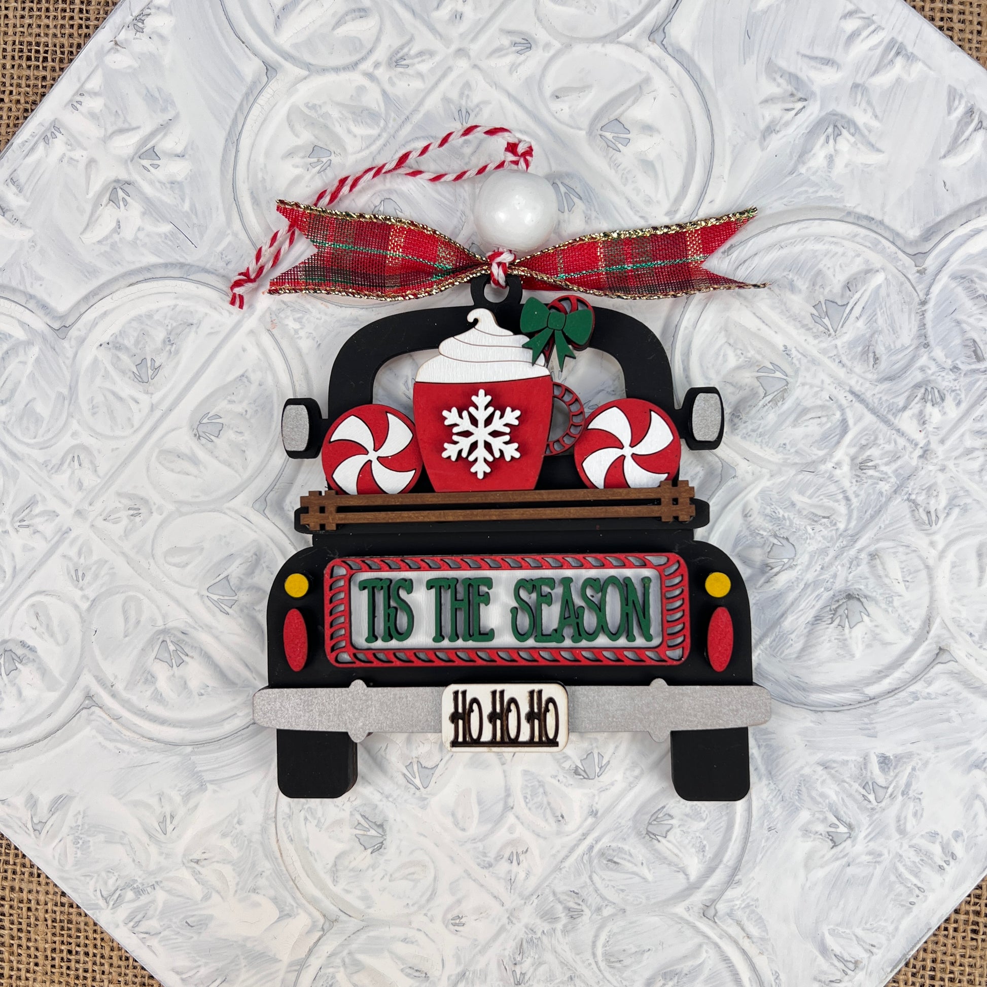 The Antique Truck Ornament from Janet's Craft Corner features a charming antique truck carrying a red mug adorned with a snowflake and candy canes, alongside a sign that reads "Tis the Season." The truck's license plate is whimsically marked "Ho Ho Ho." This ornament comes complete with a red plaid bow and red and white twine for easy hanging, making it an ideal addition to your Christmas décor.