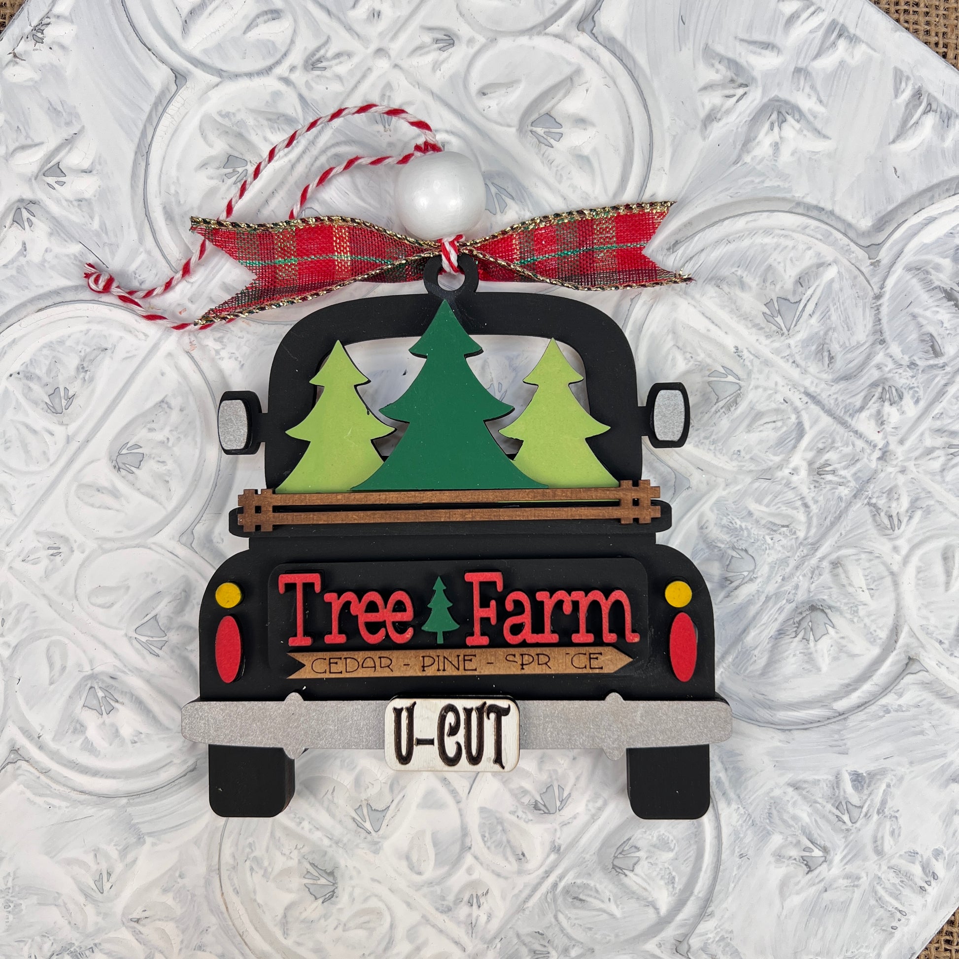 The Antique Truck Ornament from Janet's Craft Corner features a charming black truck carrying three green trees, with "Tree Farm" elegantly displayed on the tailgate. It comes adorned with a festive red and green plaid bow and red and white twine at the top, along with "U-Cut" written at the bottom. This piece is perfect for Christmas décor enthusiasts!