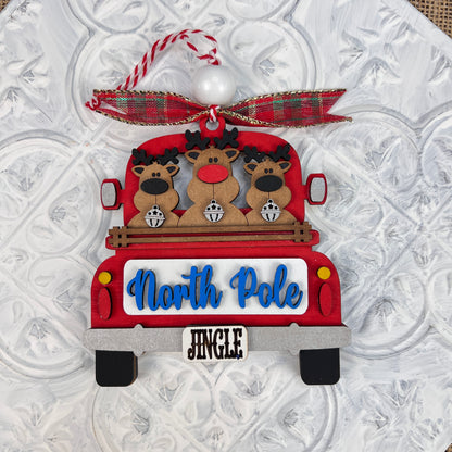 A delightful ornament from Janet's Craft Corner, the Antique Truck Ornament is part of their DIY Decoration Kits. It showcases three reindeer in an antique red truck with a "North Pole" sign and a "Jingle" license plate, adorned with a red and green plaid bow on top against a textured white background. Perfect for adding charm to your Christmas décor collection.