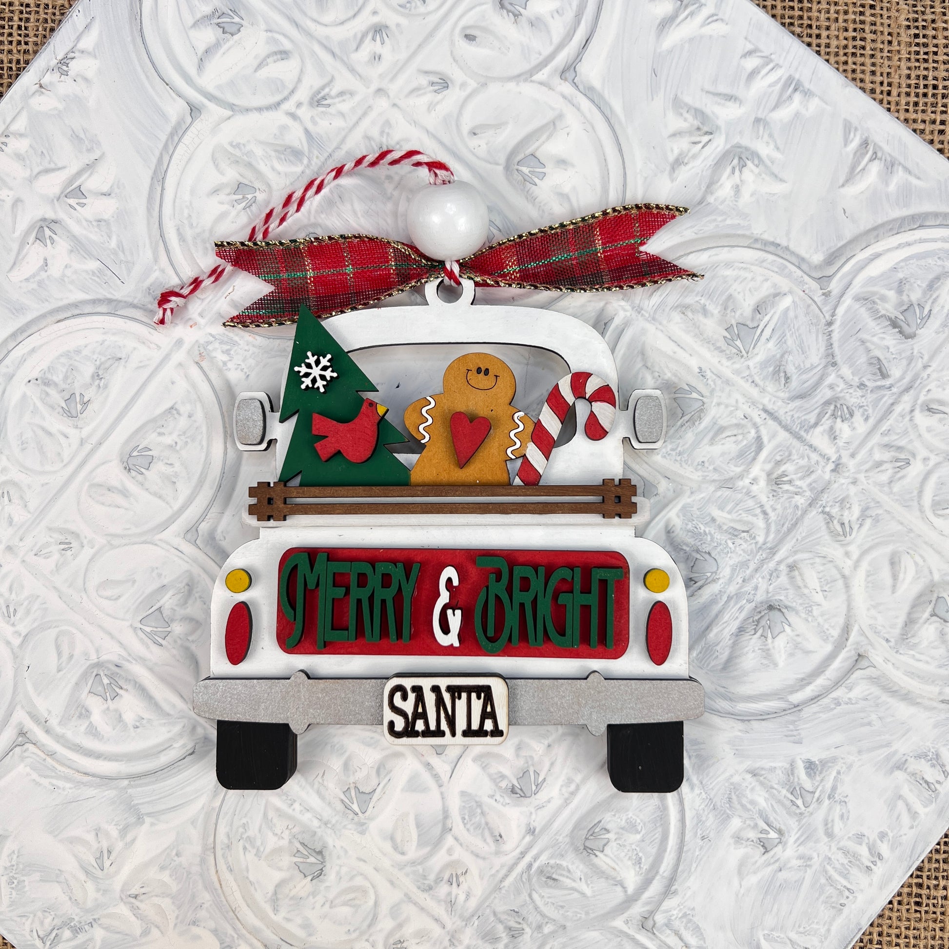 The Antique Truck Ornament from Janet's Craft Corner is a delightful piece of Christmas décor, showcasing a "Merry & Bright" sign at the back. It features a Christmas tree, a cheerful gingerbread figure, and a candy cane, all enhanced with a red plaid ribbon and white bead to highlight its vintage charm.