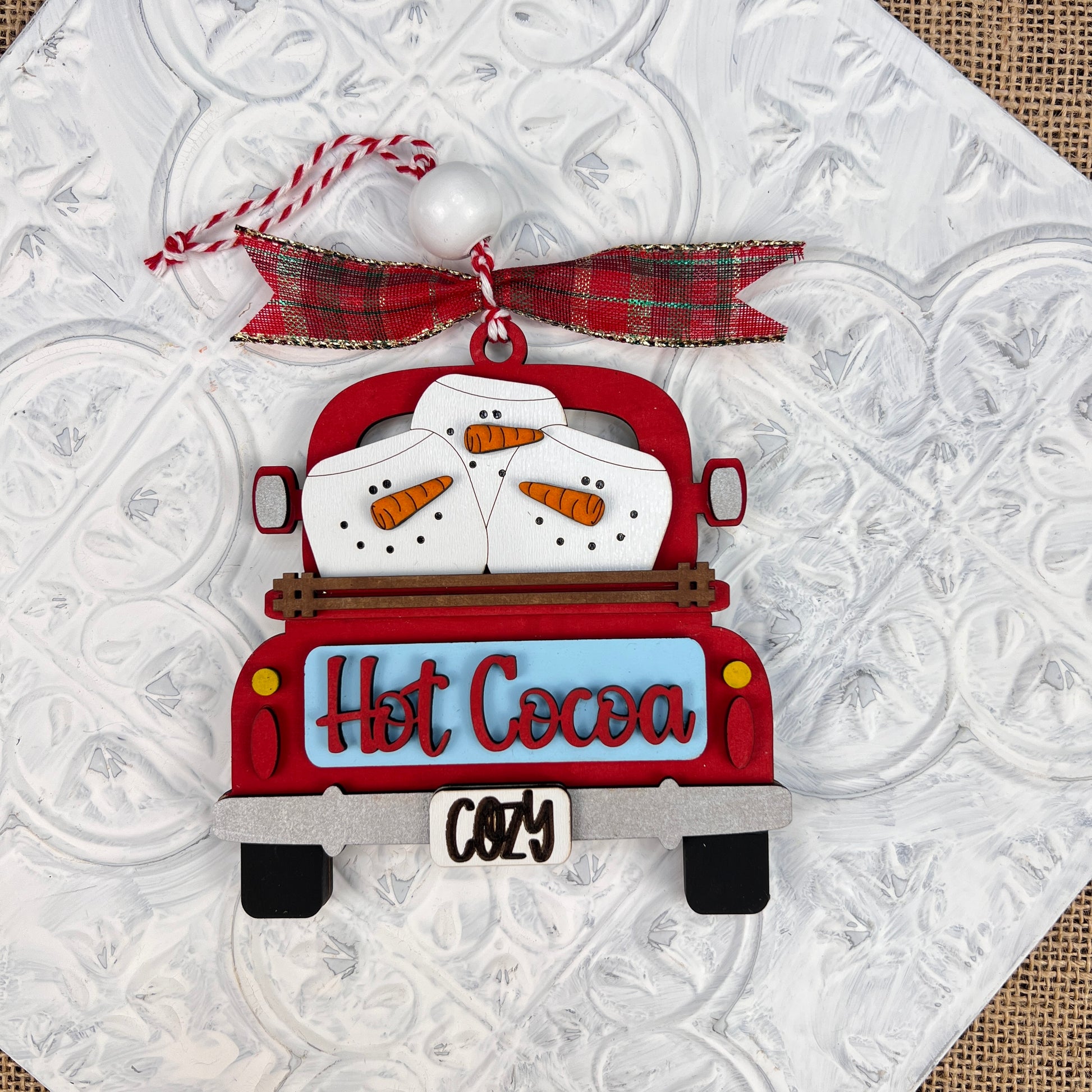 Experience nostalgic holiday charm with the Antique Truck Ornament from Janet's Craft Corner. This festive DIY decoration kit features an antique red truck carrying snowman marshmallows in the back, playfully inscribed with "Hot Cocoa" and sporting a "Cozy" license plate. The ornament is adorned with a red plaid bow and hangs beautifully against a textured white background, perfect for your Christmas décor.