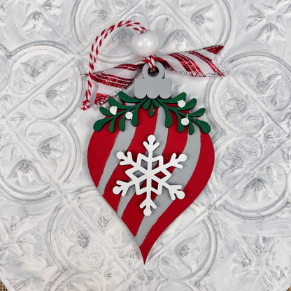 The Mistletoe Ornaments DIY Decor Kit by Janet's Craft Corner features red and silver teardrop ornaments with a white snowflake and green foliage, hanging from red and white twine. A red striped ribbon and white bead add charm to the textured backdrop.