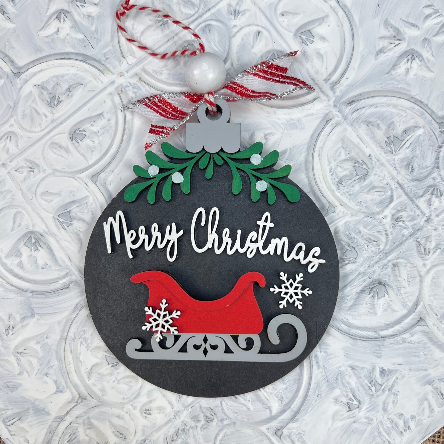 A DIY craft kit from Janet's Craft Corner includes round Christmas ornaments featuring white "Merry Christmas" script, a red sled, mistletoe with two white snowflakes, green leaves, and a red-striped bow on a textured white backdrop. Ideal for personalized home décor enthusiasts.