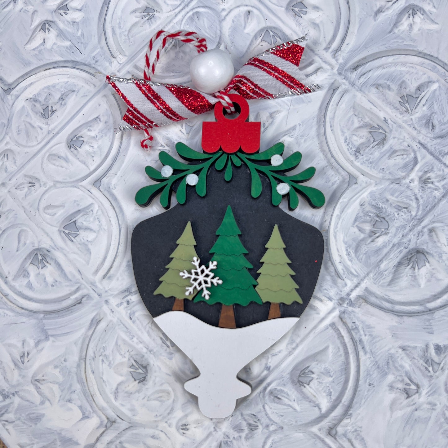 The Mistletoe Ornaments DIY Decor Kit by Janet's Craft Corner is a set of 6 charming Christmas baubles decorated with green pine trees, snowflakes, and topped with festive red and green sprigs. They feature a red and white ribbon and bead on a textured white background, ideal for personalized home décor.