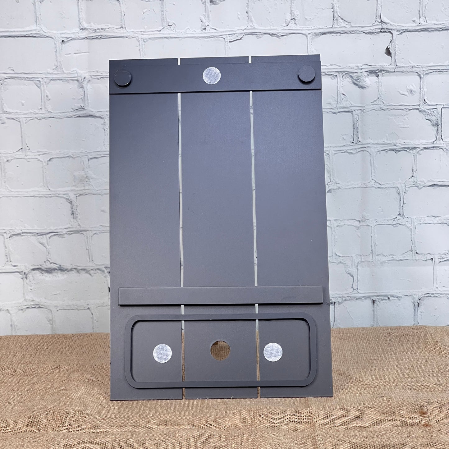 A gray, rectangular Pallet Interchangeable Base with Insert by Janet's Craft Corner, featuring circular pads, a slot near the bottom, and a slanted design, is shown on burlap before a white brick wall.