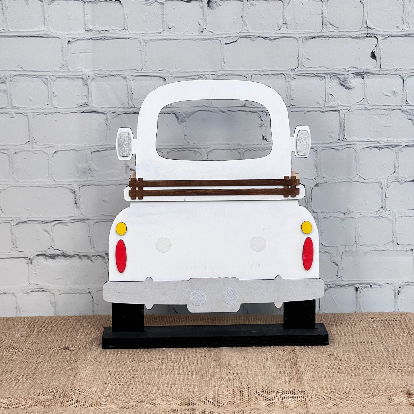 The Antique Truck Interchangeable Base - Painted from Janet's Craft Corner is a wooden white vintage truck cutout with red and yellow taillights on a black stand, ideal for mantel or craft displays.