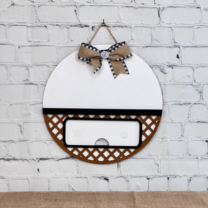 Janet's Craft Corner offers a DIY kit featuring an interchangeable base with a white, brown lattice round wall piece against a white-brick backdrop. It has black and white plaid ribbons, a button on top, and a central rectangular cutout for added charm.