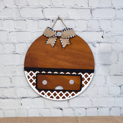 This circular brown wooden wall decoration from Janet's Craft Corner features a black and white checkered bow on a lattice pattern against a white brick background. It includes an interchangeable base with insert, perfect for DIY customization by home décor enthusiasts.