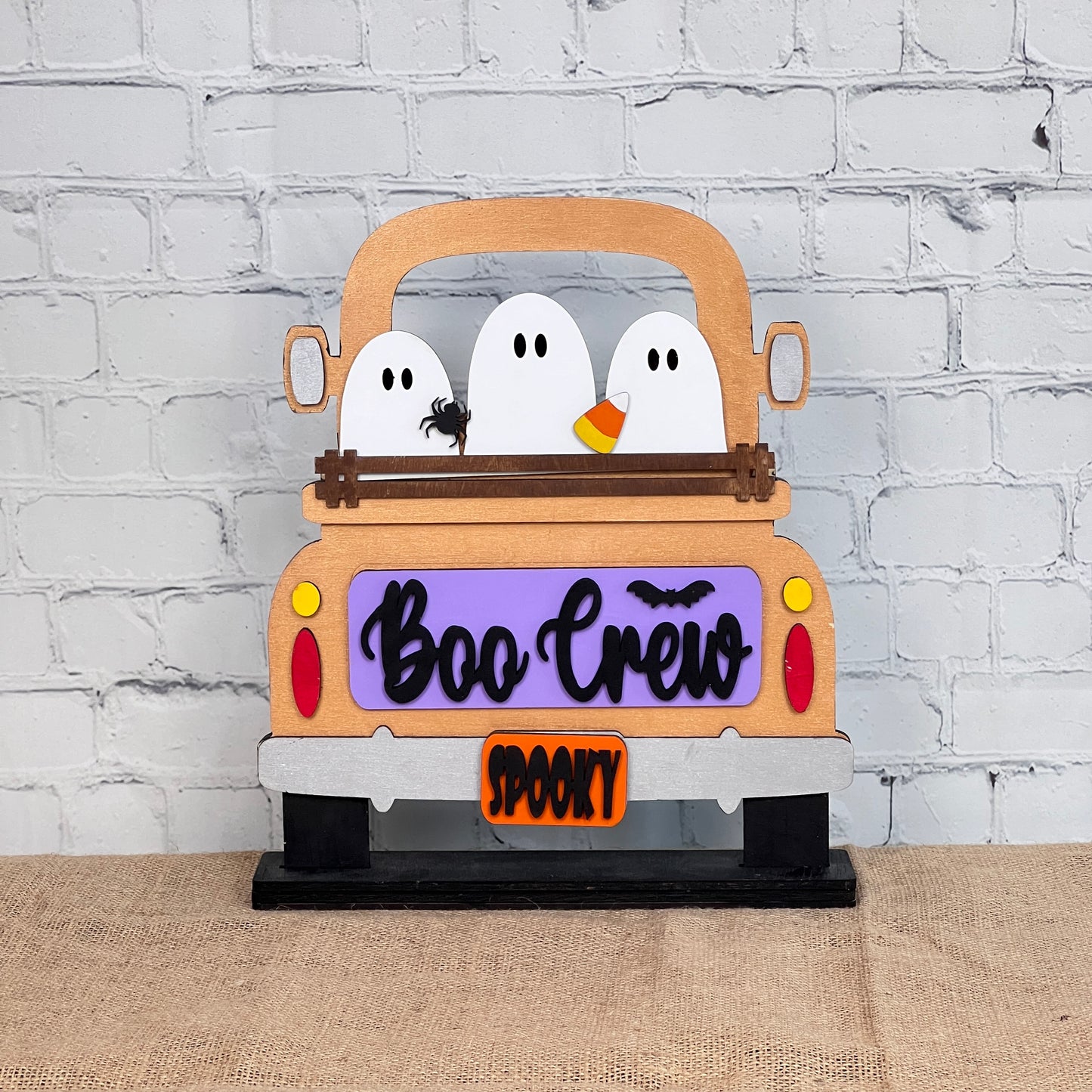 Janet's Craft Corner offers the Boo Crew Interchangeable Insert, a home decor set featuring three cartoon ghosts in a colorful pickup with charming sidekicks—a bat and spider—all set against a white brick wall.