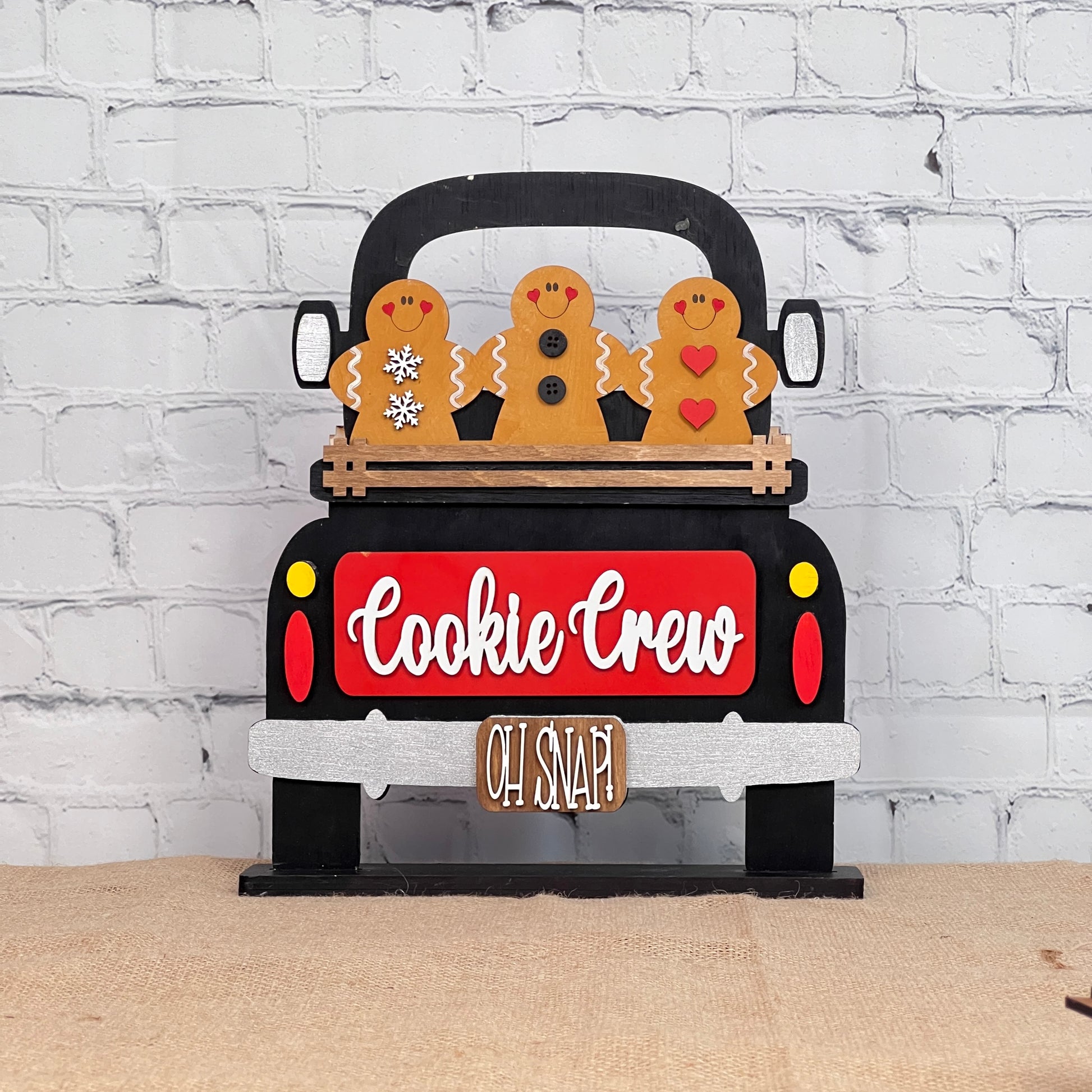 The Cookie Crew Interchangeable Insert DIY kit from Janet's Craft Corner features a black and red truck with "Cookie Crew" on its tailgate, three gingerbread men in the bed, and the phrase "Oh Snap!" dangling below. It's a delightful craft that stands out against a white brick wall.