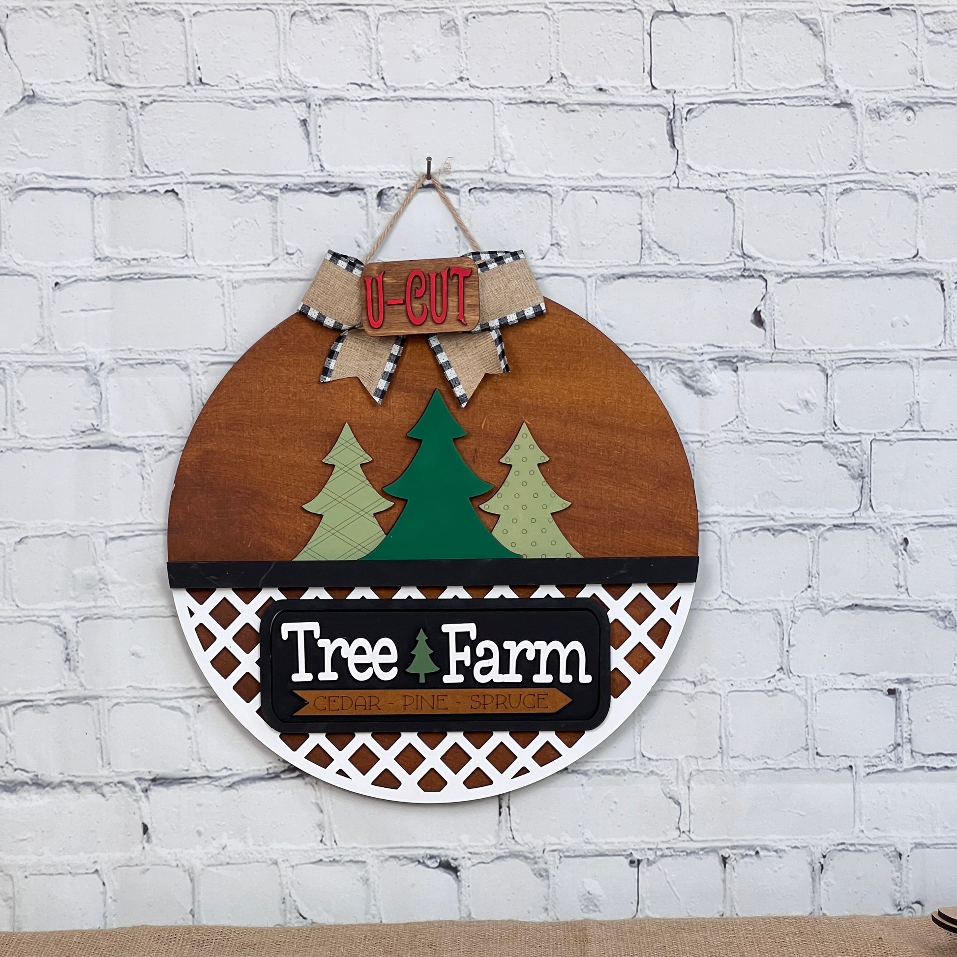 A circular wooden sign from Janet's Craft Corner's Tree Farm Interchangeable Insert DIY kit hangs on a white brick wall, featuring three green pine trees with "Tree Farm" text above "Cedar Pine Spruce." A festive bow labeled "U-CUT" completes this perfect holiday decoration.