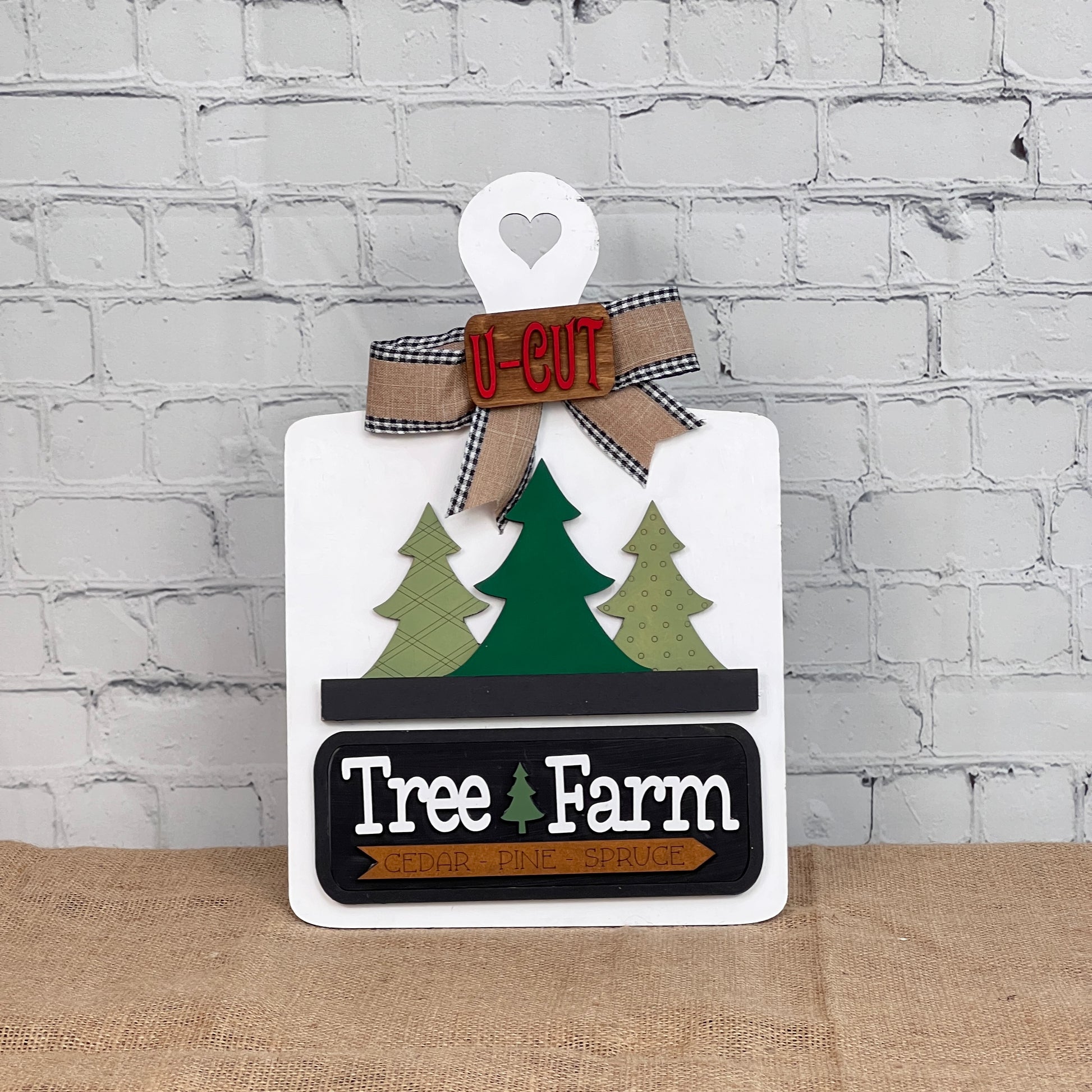 Janet's Craft Corner's Tree Farm Interchangeable Insert DIY home decor kit is a charming holiday sign resembling a cutting board with a heart cutout. It features two green trees, "Tree Farm," "Cedar Pine Spruce," and an "U-CUT" ribbon set against a white brick wall backdrop.