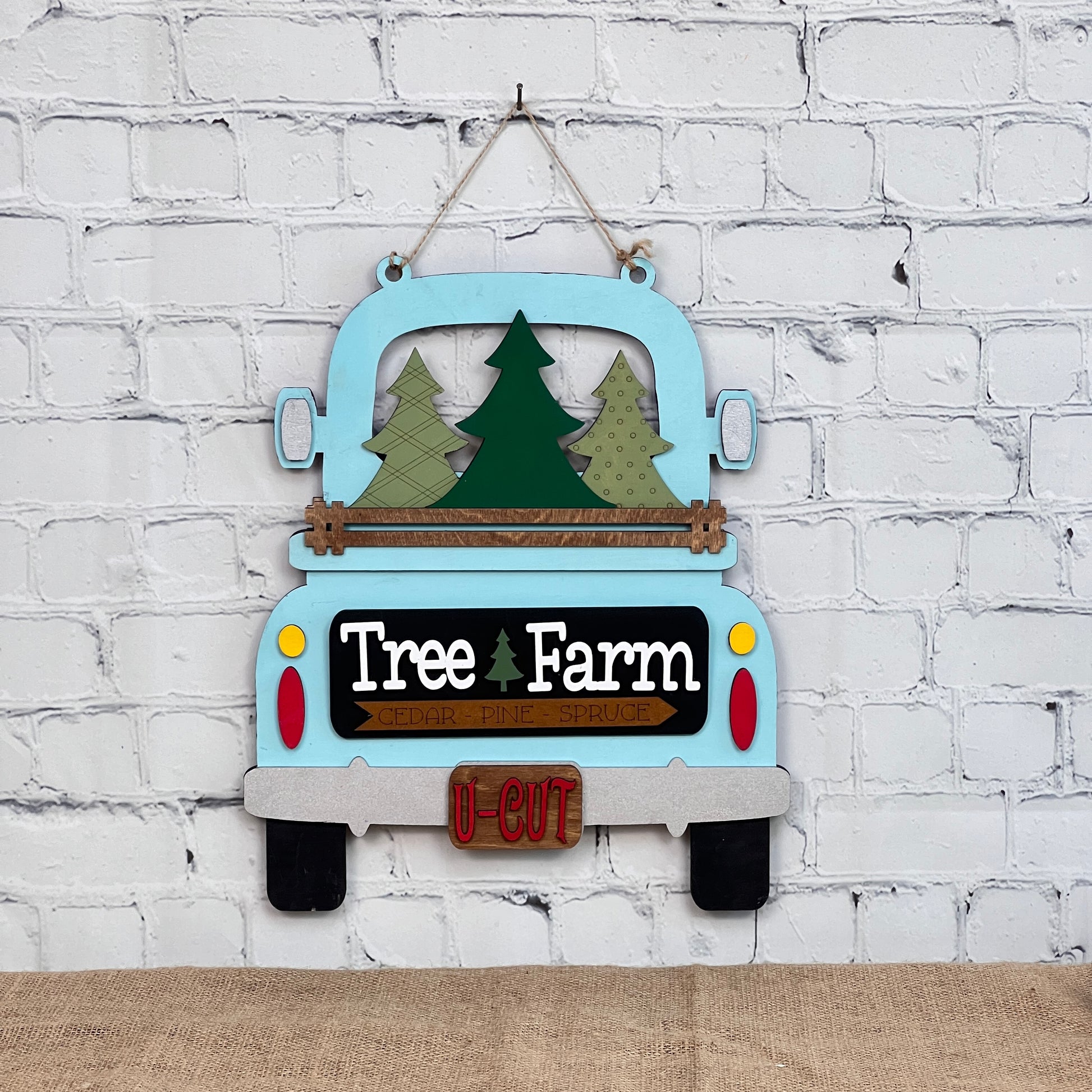 The Tree Farm Interchangeable Insert from Janet's Craft Corner is a DIY home decor kit featuring a vintage blue truck wall hanging with green tree cutouts, "Tree Farm" lettering, and "U-Cut" on a red license plate, perfect for enhancing holiday decorations against a white brick wall.