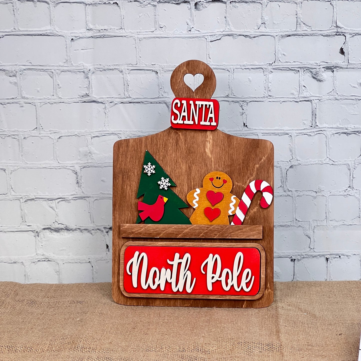 Janet's Craft Corner offers the North Pole Interchangeable Insert, a DIY seasonal home decor kit. It includes a wooden sign with a handle and heart cutout labeled "Santa," plus decorative pieces—a green tree, gingerbread man, and candy cane—with "North Pole" on a red background at the bottom.