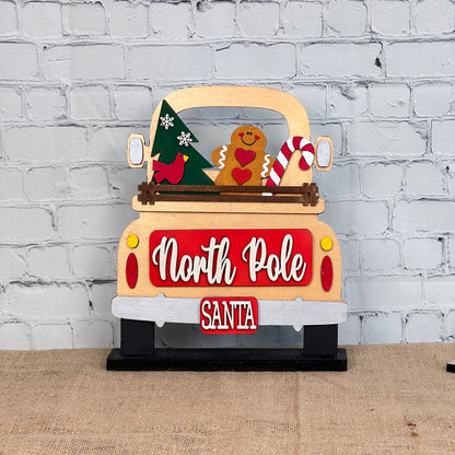 The North Pole Interchangeable Insert from Janet's Craft Corner is a festive DIY seasonal home decor kit shaped like a truck's rear, featuring "North Pole" in white script. It includes a Christmas tree, gingerbread man, candy cane in the truck bed, and "Santa" on the bumper, all displayed on burlap.