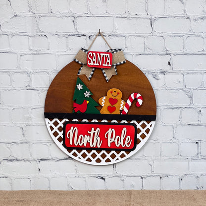 The North Pole Interchangeable Insert by Janet's Craft Corner is a DIY holiday decor kit featuring vibrant red and white details, with a gingerbread man, Christmas tree, candy cane, and red bird. It's adorned with a burlap ribbon stating "Santa" against a white brick design.