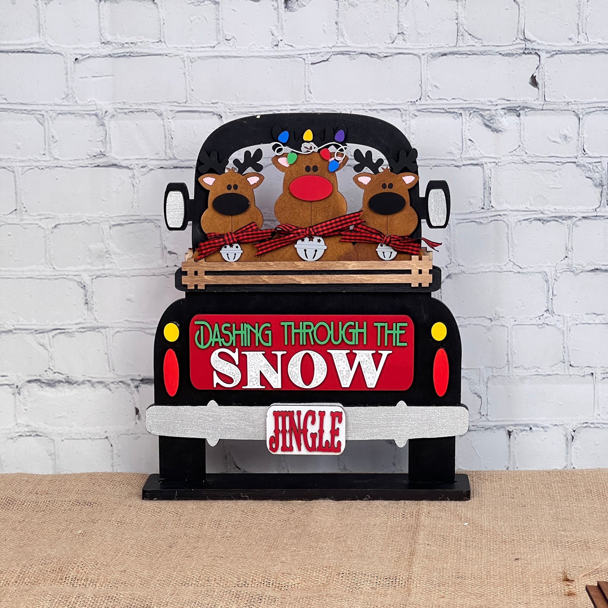 Janet's Craft Corner offers the Reindeer Interchangeable Insert, a DIY seasonal home decor kit featuring a black truck with cartoon reindeer and festive text against a white brick wall on burlap—ideal for hand-painted décor enthusiasts.