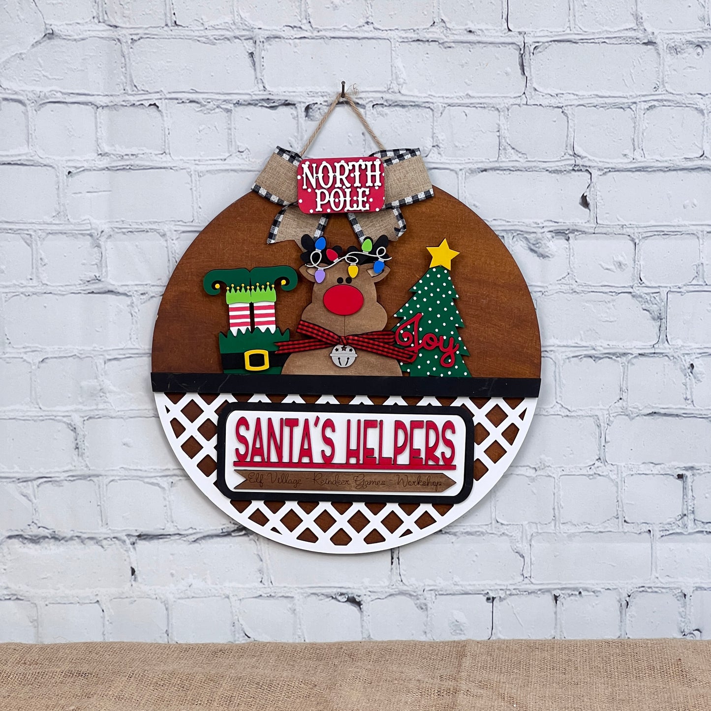 Janet's Craft Corner offers a festive wall decoration on a brick background featuring "North Pole," a light-adorned reindeer, DIY craft kit Christmas tree labeled "Joy," and an elf boot, all bundled with the "Santa's Helpers" Interchangeable Insert. Perfect for holiday decor fans!.