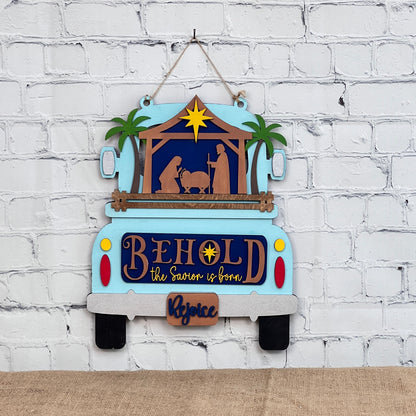 A decorative wall hanging from Janet's Craft Corner, featuring a blue truck with an interchangeable Nativity decor insert, reads "Behold the Savior is born" and "Rejoice" on the tailgate. It captures the holiday spirit beautifully against a white brick wall.