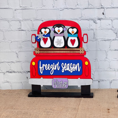 Janet's Craft Corner offers a DIY interchangeable home decor kit featuring three penguins in scarves on an antique truck. With "freezin' season" and "Winter" on the license plate, this charming piece, set against a white brick wall, adds a cozy touch to any space.