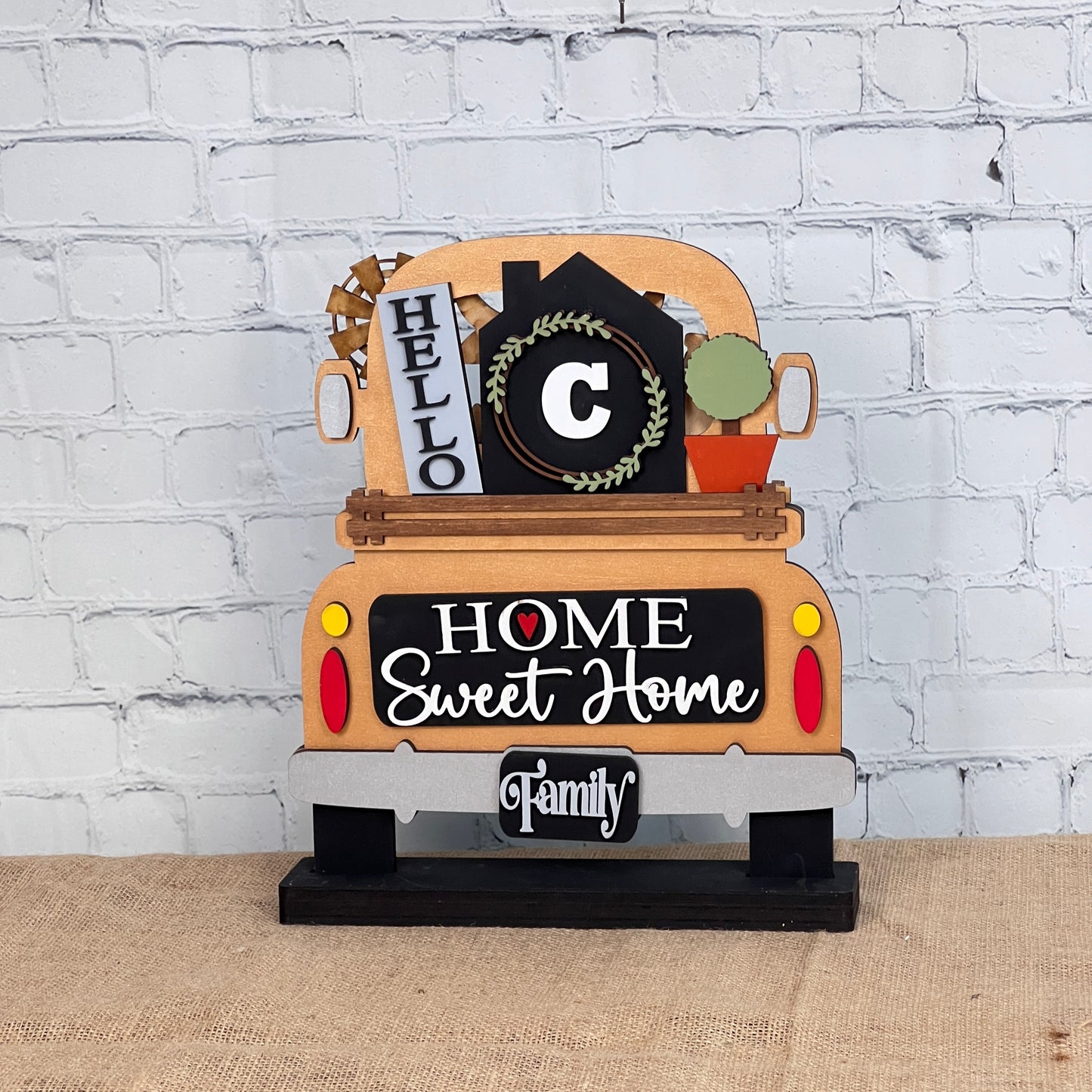 A Home Monogram Interchangeable Insert from Janet's Craft Corner, featuring phrases like "Home Sweet Home" and "Family" with a wreath and "Hello," sits decoratively on burlap against a brick wall.