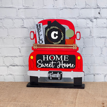 A charming red truck-shaped sign from Janet's Craft Corner features a DIY home decor kit with an interchangeable "C" monogrammed house and wreath, a "Hello" sign, and a tailgate message reading "Home Sweet Home" and "Family," all set on burlap.