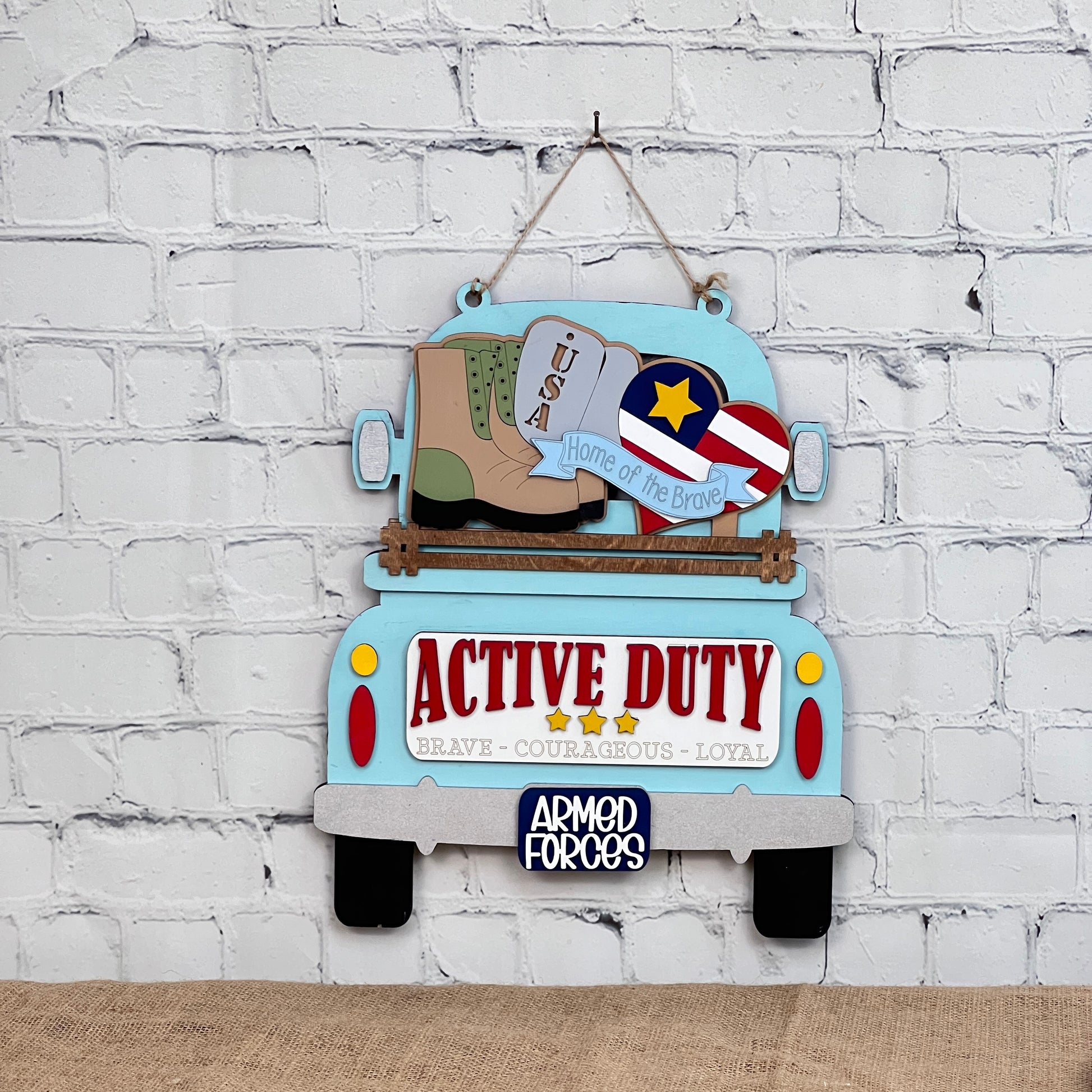 Janet's Craft Corner offers a Military Interchangeable Insert DIY kit featuring a blue vintage truck sign with "Active Duty" on the tailgate, military boots, USA dog tags, and a star emblem. Ideal for home decor against a white brick wall.