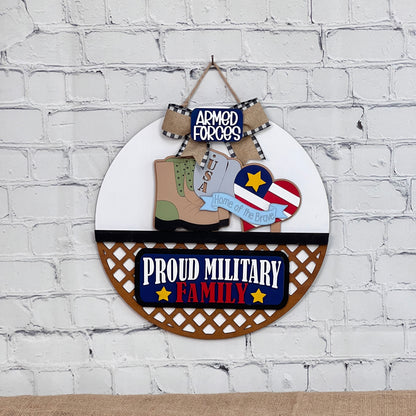 A DIY home decor kit by Janet's Craft Corner includes a circular insert featuring a boot, USA tag, stars and stripes with a heart, an "Armed Forces" plaque, and "Proud Military Family" at the bottom. It is accented with a black and white bow on a brick background.