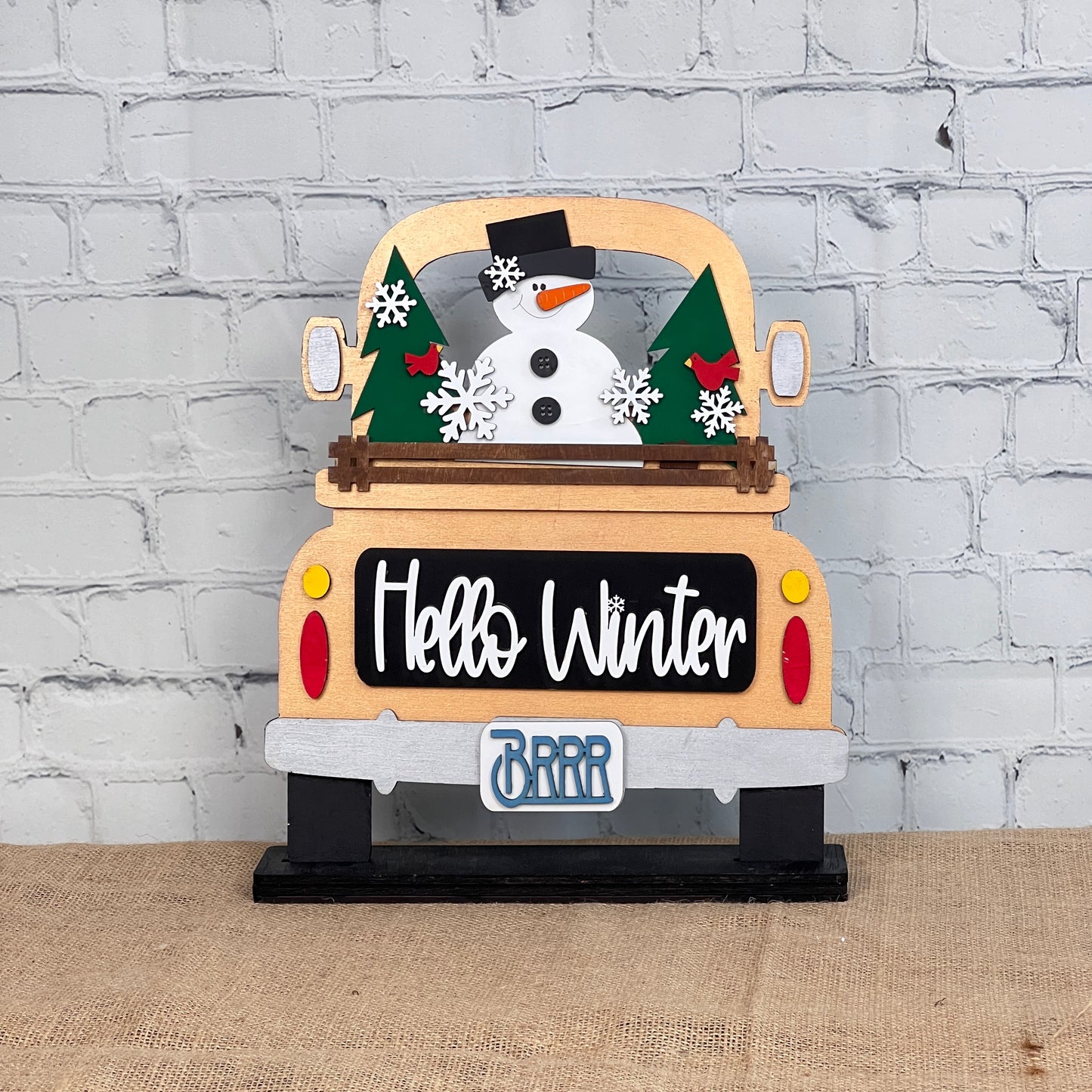 Janet's Craft Corner presents the Antique Truck Base with Insert, a DIY interchangeable home decor kit featuring a decorative wood cutout of an antique truck with a snowman, trees, and snowflakes. "Hello Winter" and "Brrr" phrases complete this charming seasonal piece against rustic brick and burlap.