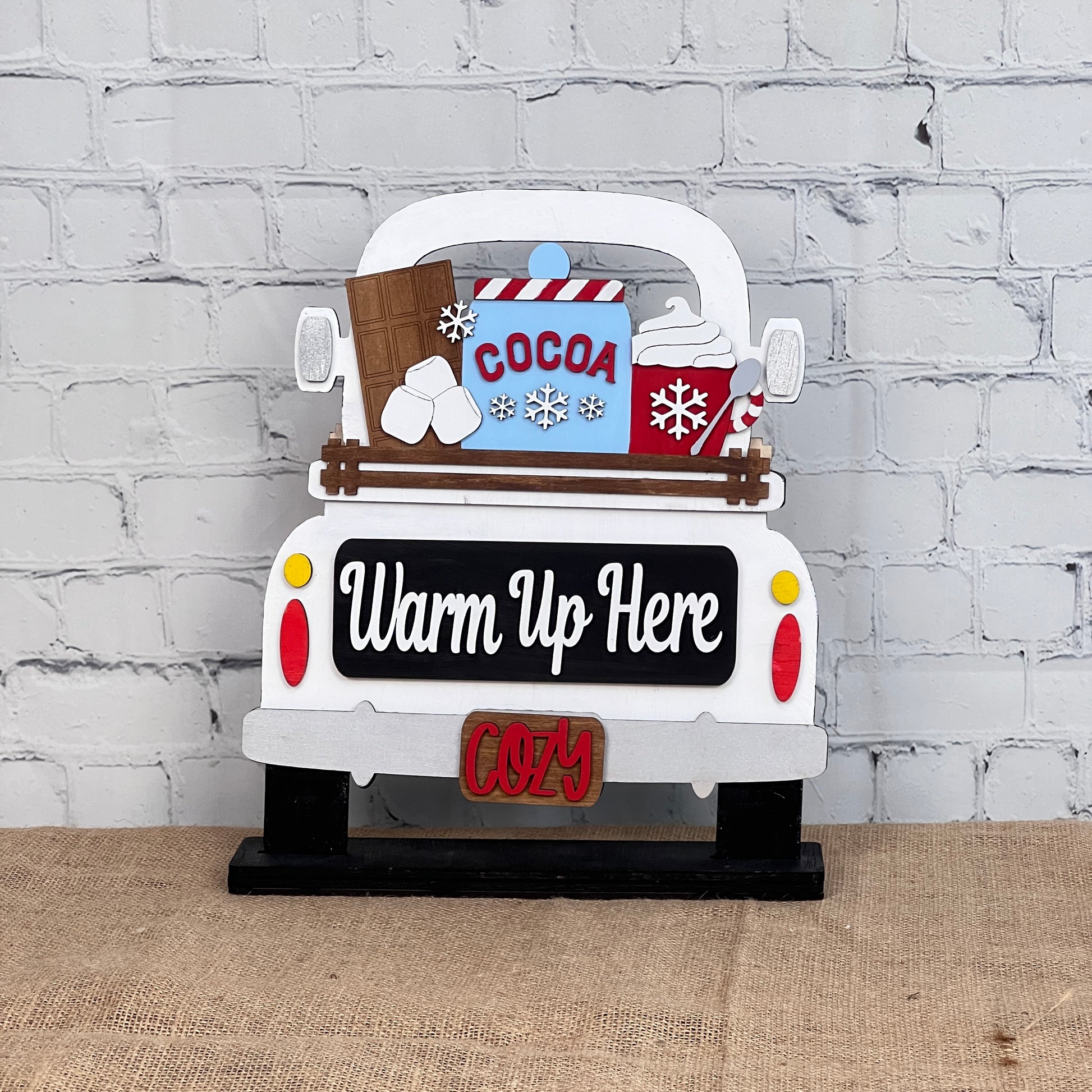 Janet's Craft Corner's Antique Truck Base with Insert is a DIY home decor kit featuring an antique truck sign against a white brick wall. It displays "Warm Up Here," with "Cocoa," marshmallows, and a mug in the bed. The license plate reads "COZY," making it perfect as interchangeable decor.