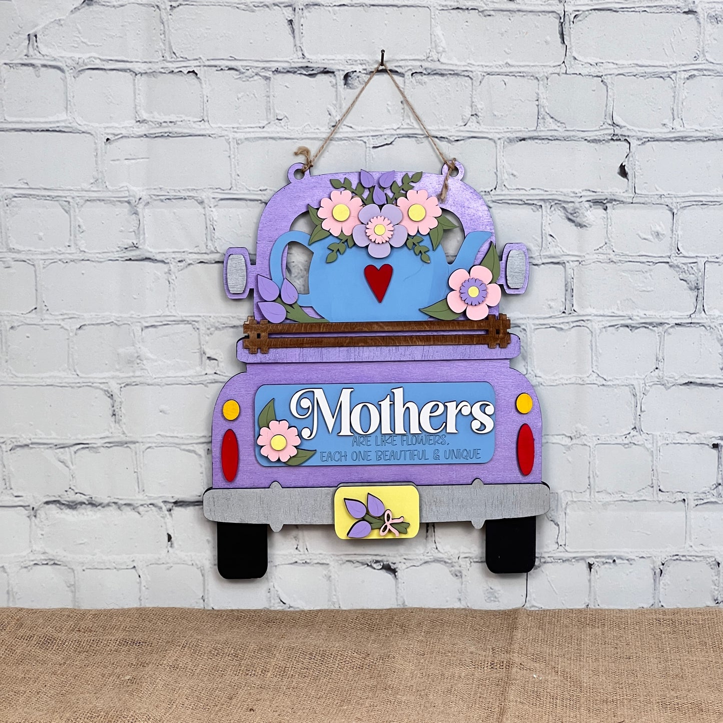 A charming decorative sign from Janet's Craft Corner features a wooden purple antique truck adorned with pink and blue flowers and "Mothers" text, perfect for hanging on a white brick wall, reminiscent of their DIY interchangeable home decor kit.