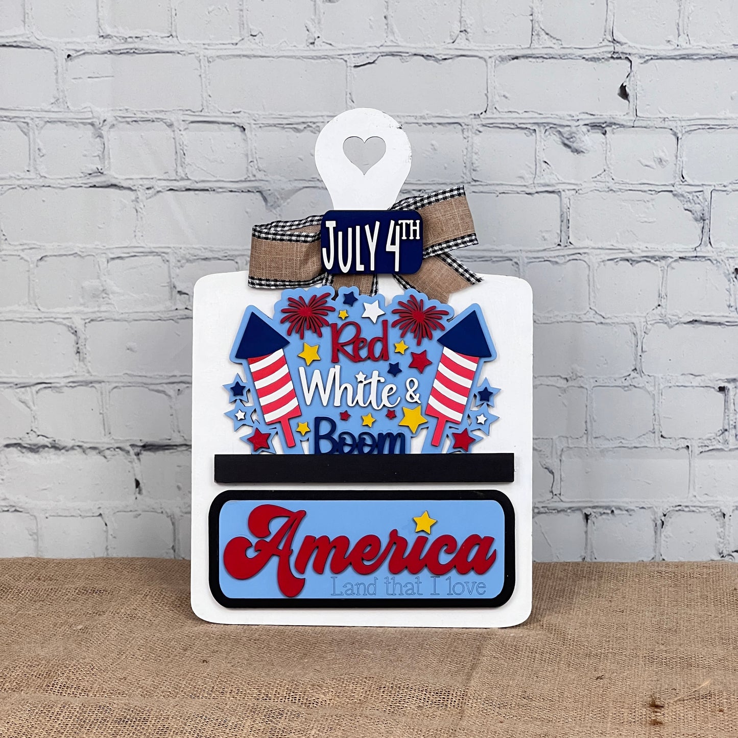 The Red White & Boom Interchangeable Insert from Janet's Craft Corner is a DIY home decor craft kit that features "July 4th," fireworks, and "Red White & Boom" in patriotic colors along with “America, Land that I Love” against a white brick wall backdrop.