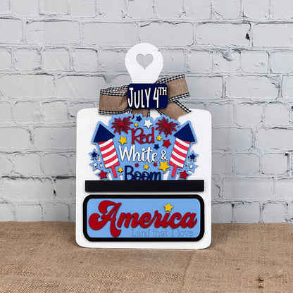 The Red White & Boom Interchangeable Insert from Janet's Craft Corner is a DIY home decor craft kit that features "July 4th," fireworks, and "Red White & Boom" in patriotic colors along with “America, Land that I Love” against a white brick wall backdrop.