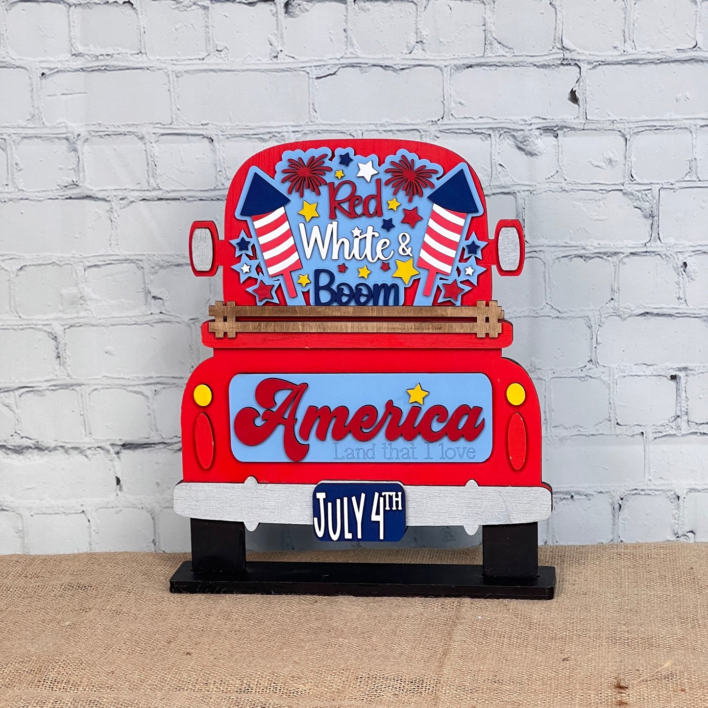 Janet's Craft Corner offers a "Red White & Boom Interchangeable Insert" featuring a patriotic red truck with fireworks and the phrases "Red White & Boom" and "America Land that I Love," plus a "July 4th" license plate, all on a white brick background—perfect for DIY seasonal home decor.