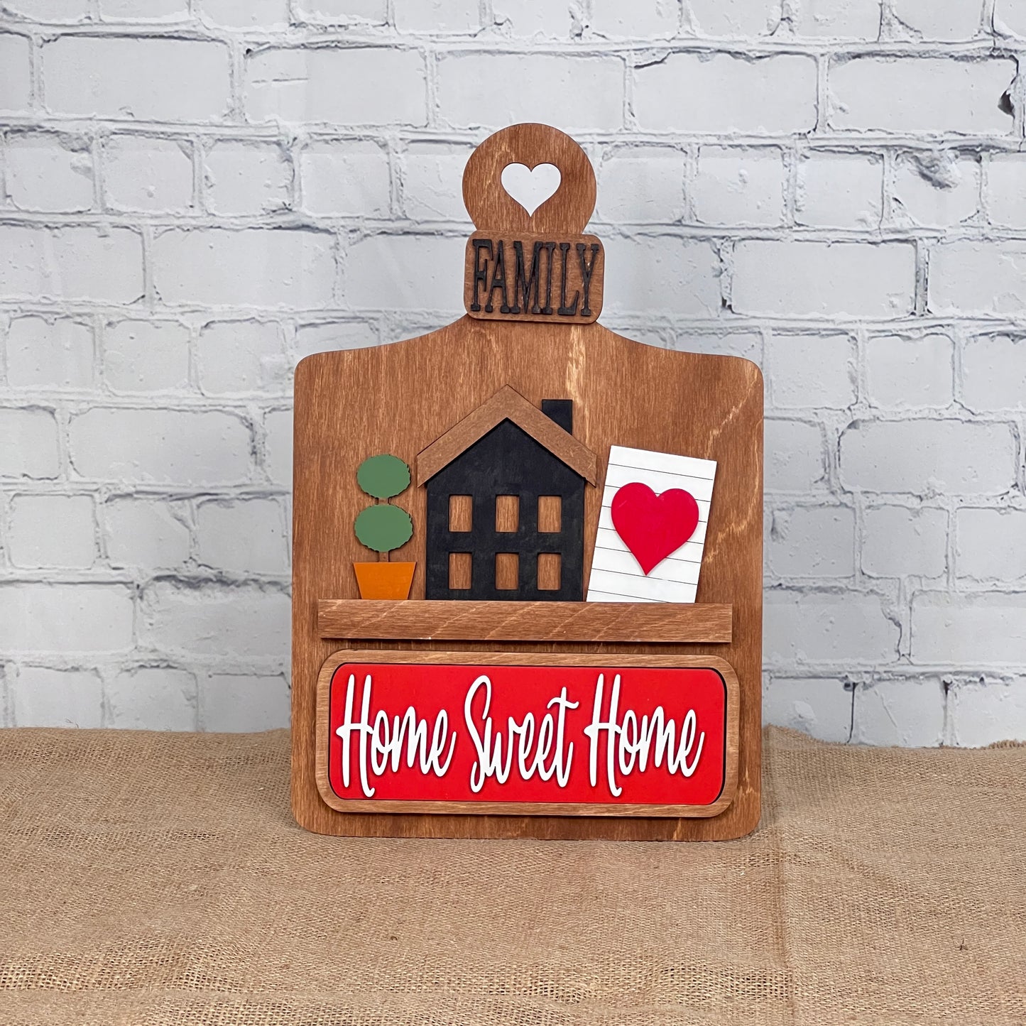 Janet's Craft Corner offers the "Home Sweet Home" Interchangeable Insert, a charming DIY home decor kit. The wooden piece features a house, potted plant, and red heart on a notecard, with "Family" on top and "Home Sweet Home" against a red background—ideal for brick wall settings.