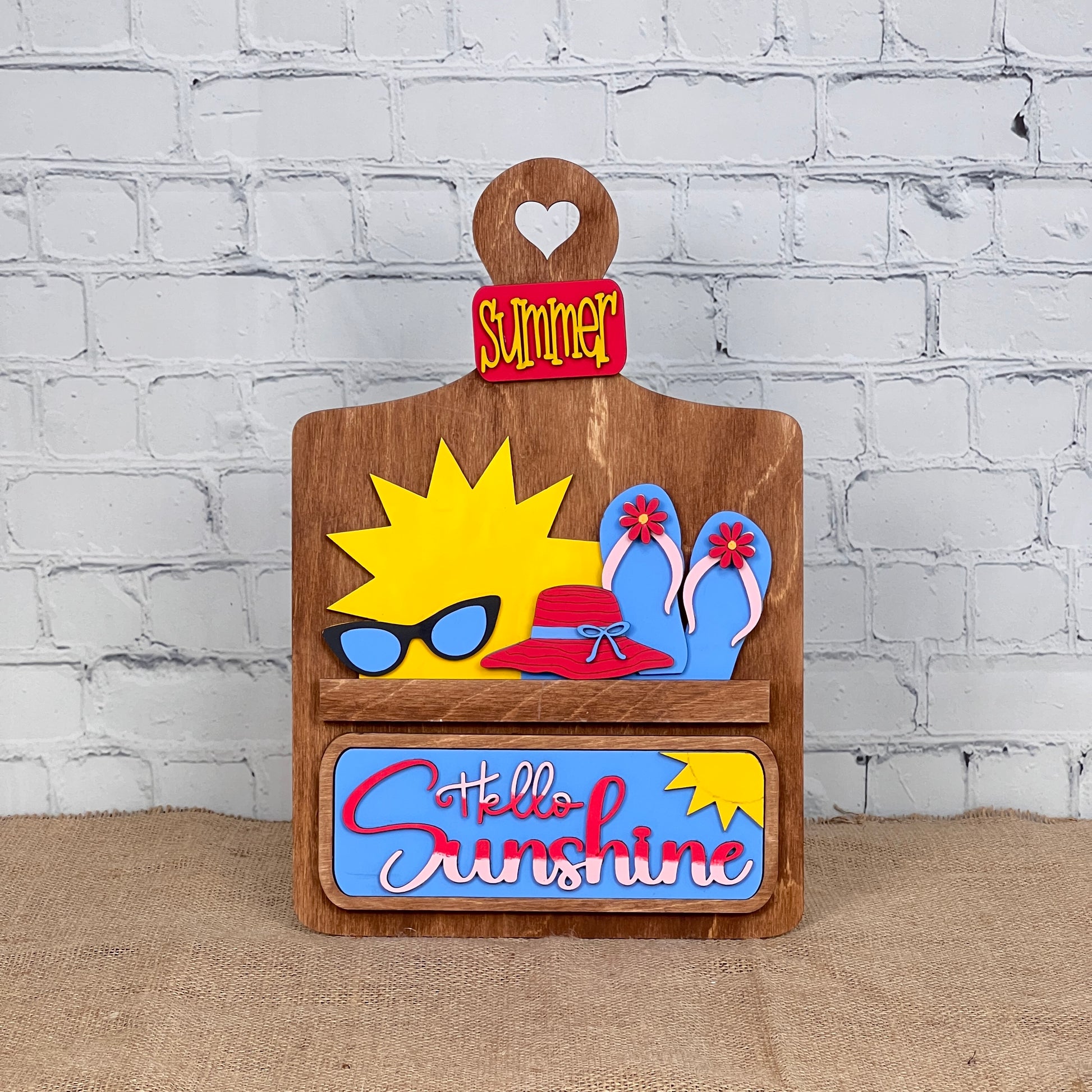 The Hello Sunshine Interchangeable Insert from Janet's Craft Corner includes a wooden decorative sign featuring the word "Summer," a heart cutout, and playful icons like sunglasses and flip-flops, perfect for adding seasonal warmth to your summer picnic against its white brick backdrop.