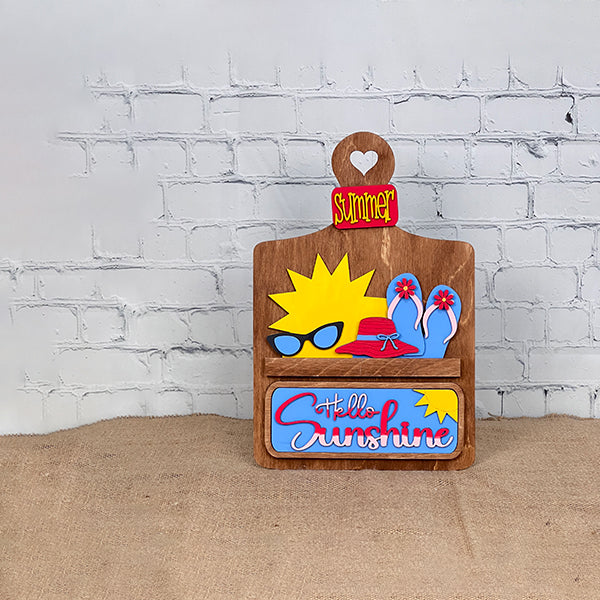 Janet's Craft Corner offers the "Hello Sunshine Interchangeable Insert" craft kit, a wooden card holder with a heart cut-out labeled "Summer." It features postcards with sun and beach motifs, all set against a white brick wall on a brown surface for seasonal home decor.